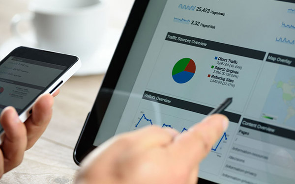 Hands hold a white cellphone and a black tablet displaying charts, graphs, and text on analytics