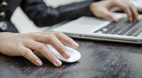 Hands using a white computer mouse and typing on a silver laptop on a black wooden desk