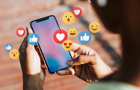 Hands holding a smartphone with social media emojis floating around the screen