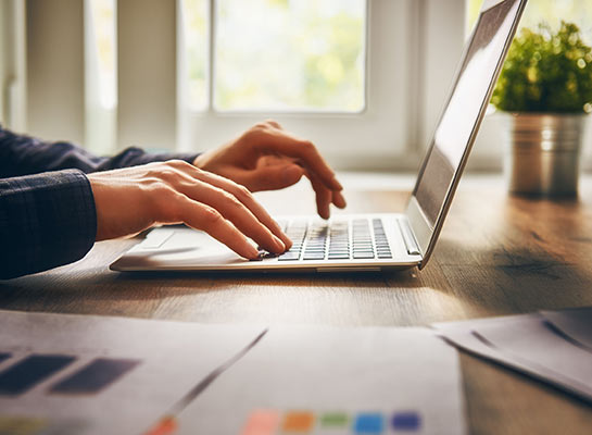 Hands typing on a silver laptop on a wooden desk with papers and a plant